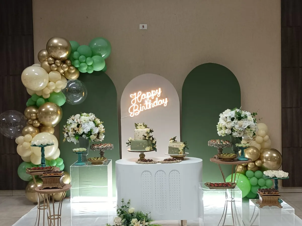 Mesa do bolo com arranjos de flores brancas, arco de balões verde e dourado e letreiro neon de aniversário