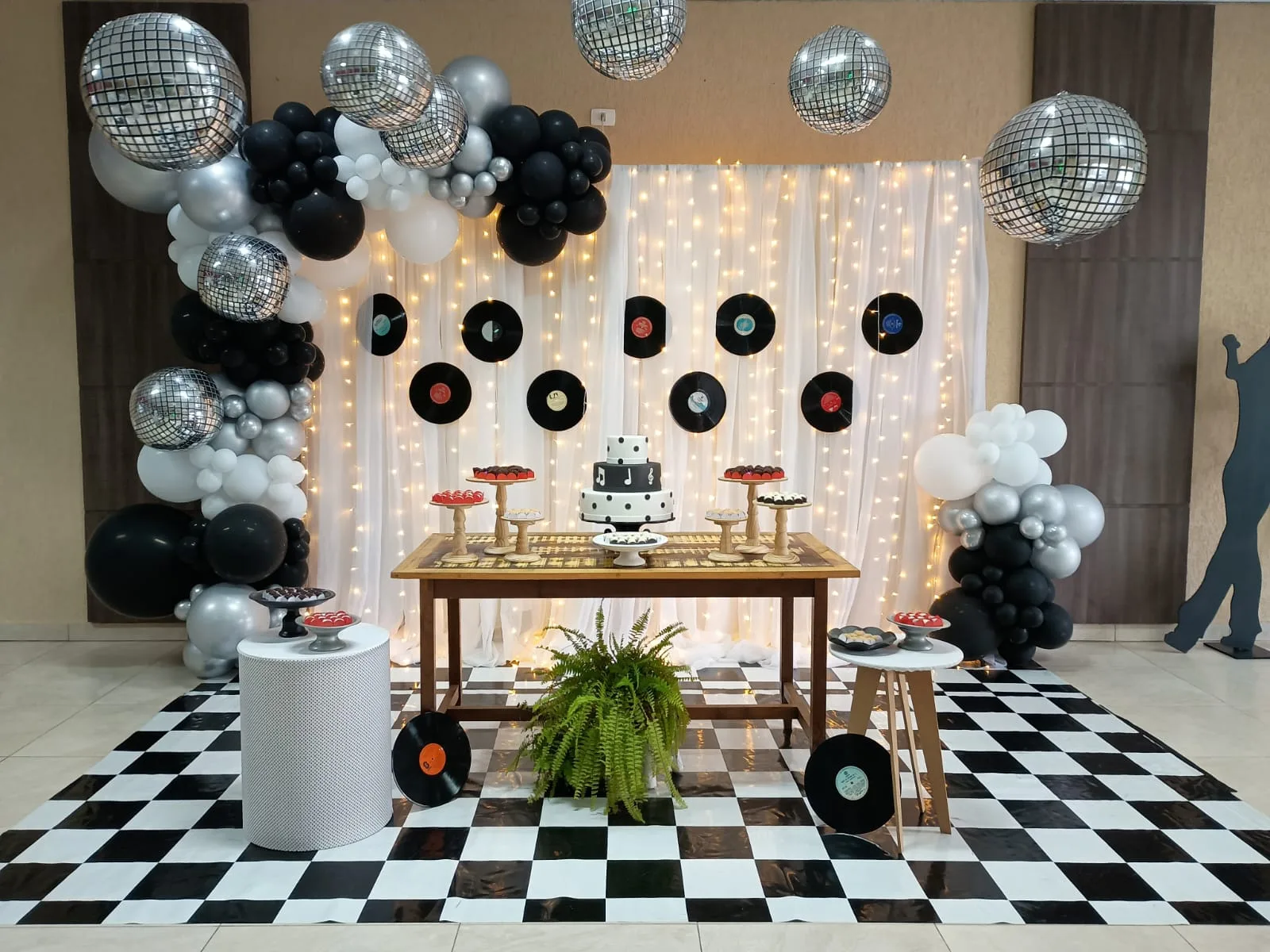 Salão decorado para festa adulta com globos espelhados, discos de vinil na parede, cortina de luzes e pista xadrez