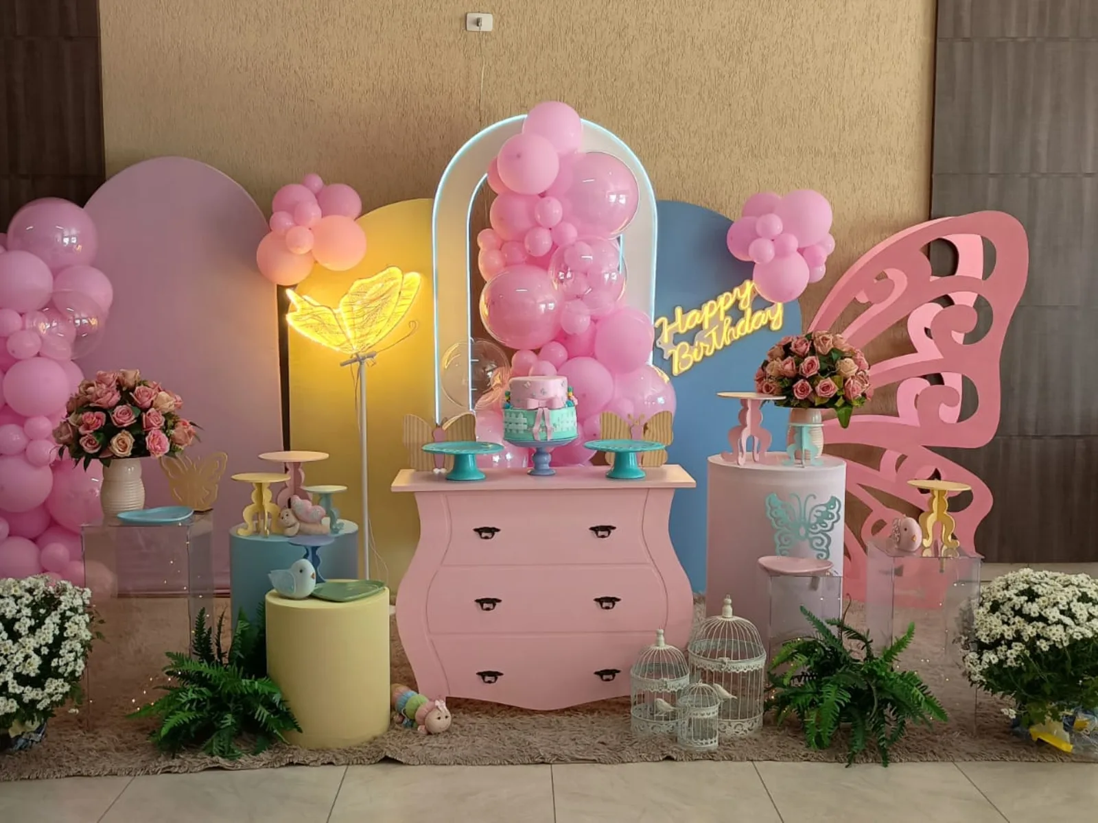 Festa infantil em tons de rosa com borboleta vazada, letreiro neon de aniversário, cômoda rosa e arranjos de flores brancas