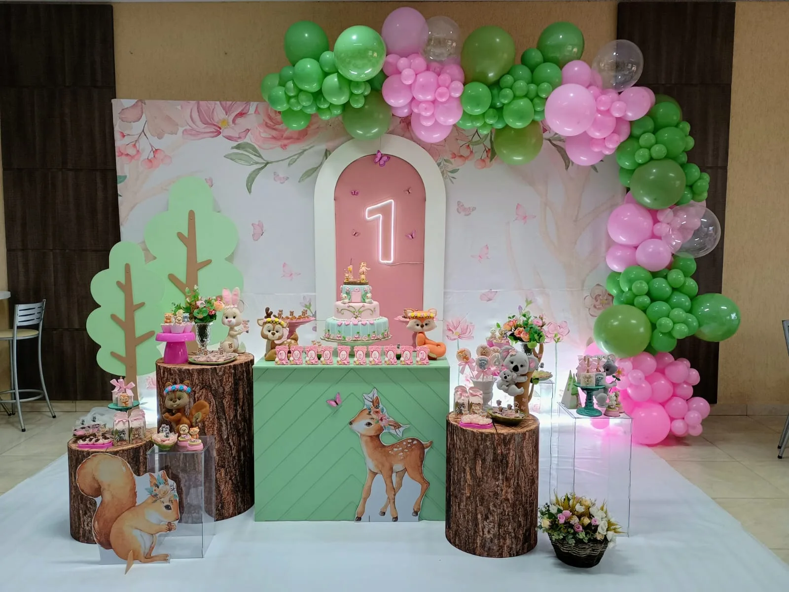 Salão decorado com cenário de bosque: corça e esquilo em troncos de madeira, arco de balões verde e rosa e bolo sobre painel floral